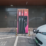 An electric vehicle charging station with a modern design is set against a dark grey building exterior. The station features three charging cables and a digital display. A white electric vehicle is parked adjacent, connected to one of the chargers.