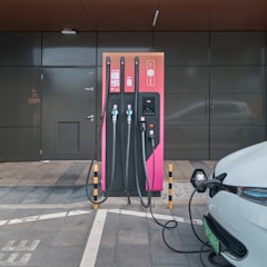 An electric vehicle charging station with a modern design is set against a dark grey building exterior. The station features three charging cables and a digital display. A white electric vehicle is parked adjacent, connected to one of the chargers.