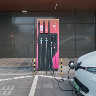 An electric vehicle charging station with a modern design is set against a dark grey building exterior. The station features three charging cables and a digital display. A white electric vehicle is parked adjacent, connected to one of the chargers.