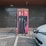 An electric vehicle charging station with a modern design is set against a dark grey building exterior. The station features three charging cables and a digital display. A white electric vehicle is parked adjacent, connected to one of the chargers.