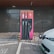 An electric vehicle charging station with a modern design is set against a dark grey building exterior. The station features three charging cables and a digital display. A white electric vehicle is parked adjacent, connected to one of the chargers.