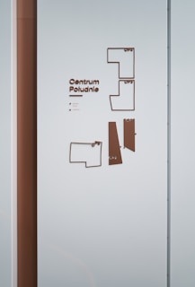 A wall features a minimalist directory map with the text 'Centrum Południe'. The map consists of outlined shapes representing different sections and floors, each labeled accordingly. The color scheme mainly includes shades of brown and light blue. The wall also shows a vertical brown panel on the left side.