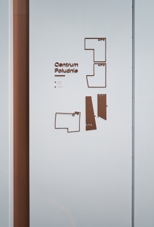 A wall features a minimalist directory map with the text 'Centrum Południe'. The map consists of outlined shapes representing different sections and floors, each labeled accordingly. The color scheme mainly includes shades of brown and light blue. The wall also shows a vertical brown panel on the left side.