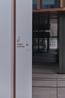 A modern building entrance featuring a clean, white wall with signage that instructs to clean up after one's dog. The wall is juxtaposed against a series of reflective windows and dark panels, creating a contrast in colors and textures. The interior appears to have a tiled floor leading into a shadowed corridor.