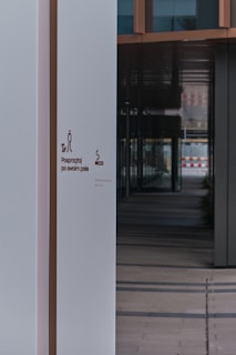 A modern building entrance featuring a clean, white wall with signage that instructs to clean up after one's dog. The wall is juxtaposed against a series of reflective windows and dark panels, creating a contrast in colors and textures. The interior appears to have a tiled floor leading into a shadowed corridor.