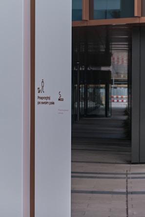 A modern building entrance featuring a clean, white wall with signage that instructs to clean up after one's dog. The wall is juxtaposed against a series of reflective windows and dark panels, creating a contrast in colors and textures. The interior appears to have a tiled floor leading into a shadowed corridor.