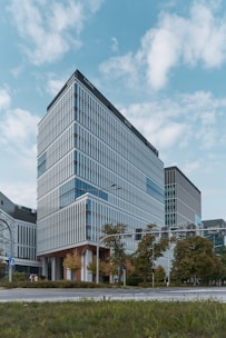 A modern office building with a sleek, rectangular design, featuring large glass windows and uniform vertical lines. The structure is surrounded by greenery, with trees lining the street in front of it. The sky is partially cloudy, providing a soft backdrop to the scene.