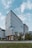 A modern office building with a sleek, rectangular design, featuring large glass windows and uniform vertical lines. The structure is surrounded by greenery, with trees lining the street in front of it. The sky is partially cloudy, providing a soft backdrop to the scene.