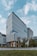 A modern office building with a sleek, rectangular design, featuring large glass windows and uniform vertical lines. The structure is surrounded by greenery, with trees lining the street in front of it. The sky is partially cloudy, providing a soft backdrop to the scene.