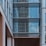 A modern building exterior featuring large glass windows with reflections of the sky and surrounding structures. The facade is a mix of straight lines and geometric shapes, primarily composed of steel and glass with some sections of beige and white materials.
