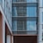 A modern building exterior featuring large glass windows with reflections of the sky and surrounding structures. The facade is a mix of straight lines and geometric shapes, primarily composed of steel and glass with some sections of beige and white materials.