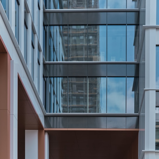 A modern building exterior featuring large glass windows with reflections of the sky and surrounding structures. The facade is a mix of straight lines and geometric shapes, primarily composed of steel and glass with some sections of beige and white materials.