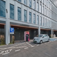 A modern electric vehicle is parked and charging at a charging station outside a sleek, glass-panelled office building. The area is marked with designated parking lines and a sign indicating an electric vehicle charging zone. Tall grasses and ornamental plants line the pavement, adding a touch of greenery to the urban setting.