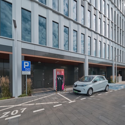 A modern electric vehicle is parked and charging at a charging station outside a sleek, glass-panelled office building. The area is marked with designated parking lines and a sign indicating an electric vehicle charging zone. Tall grasses and ornamental plants line the pavement, adding a touch of greenery to the urban setting.
