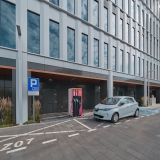 A modern electric vehicle is parked and charging at a charging station outside a sleek, glass-panelled office building. The area is marked with designated parking lines and a sign indicating an electric vehicle charging zone. Tall grasses and ornamental plants line the pavement, adding a touch of greenery to the urban setting.