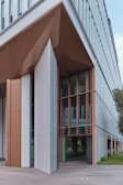 A modern building features angular, geometric architecture with large glass windows and panels. The structure is a mix of white and brown tones with metallic accents, creating a sleek, contemporary look. Greenery is visible beside the building, adding a touch of nature.