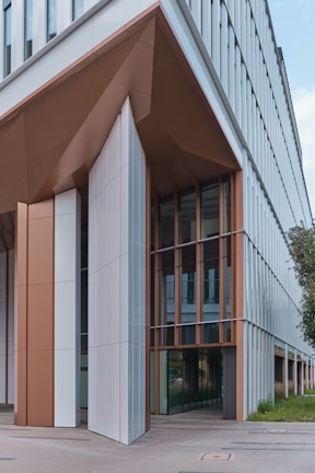 A modern building features angular, geometric architecture with large glass windows and panels. The structure is a mix of white and brown tones with metallic accents, creating a sleek, contemporary look. Greenery is visible beside the building, adding a touch of nature.
