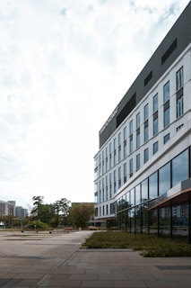 A modern office building with a sleek glass facade stretching upward, located in an urban setting. The building is surrounded by a tiled sidewalk and landscaped greenery. The sky is overcast, creating a soft lighting effect.