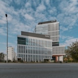 A modern, multi-story office building with a sleek design featuring large glass windows and geometric patterns. The building is set against a backdrop of a partly cloudy sky. Surroundings include a few trees and a lamppost, with other high-rise buildings visible in the background.