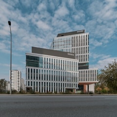 A modern, multi-story office building with a sleek design featuring large glass windows and geometric patterns. The building is set against a backdrop of a partly cloudy sky. Surroundings include a few trees and a lamppost, with other high-rise buildings visible in the background.