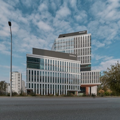 A modern, multi-story office building with a sleek design featuring large glass windows and geometric patterns. The building is set against a backdrop of a partly cloudy sky. Surroundings include a few trees and a lamppost, with other high-rise buildings visible in the background.