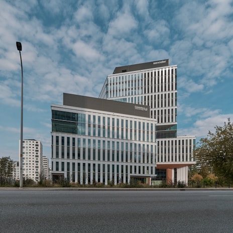 A modern, multi-story office building with a sleek design featuring large glass windows and geometric patterns. The building is set against a backdrop of a partly cloudy sky. Surroundings include a few trees and a lamppost, with other high-rise buildings visible in the background.