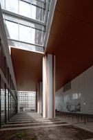 A modern architectural space featuring large vertical columns, a high ceiling with glass panels that allow natural light to stream in, and a combination of white and brown elements. The area includes wall art and some benches, with a blend of indoor and outdoor elements visible through glass walls.