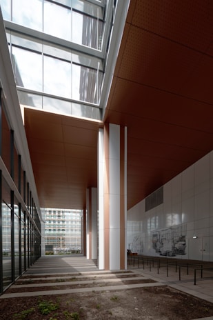 A modern architectural space featuring large vertical columns, a high ceiling with glass panels that allow natural light to stream in, and a combination of white and brown elements. The area includes wall art and some benches, with a blend of indoor and outdoor elements visible through glass walls.