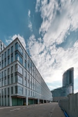 Modern architectural scene featuring a large office building with glass windows and a curving high-rise structure in the background. The sky is partly cloudy with soft, diffused light creating a calming atmosphere.