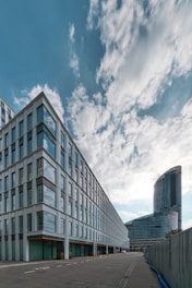 Modern architectural scene featuring a large office building with glass windows and a curving high-rise structure in the background. The sky is partly cloudy with soft, diffused light creating a calming atmosphere.