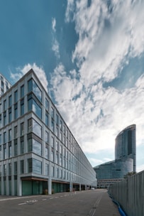 Modern architectural scene featuring a large office building with glass windows and a curving high-rise structure in the background. The sky is partly cloudy with soft, diffused light creating a calming atmosphere.