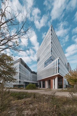 A modern architectural building with a sharp, angular design stands against a bright blue sky with scattered clouds. The structure is mostly glass and white panels, exhibiting a sleek, contemporary style. The foreground has rough terrain with some greenery and trees, suggesting an urban environment.