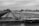 A black and white image featuring a large river barge laden with gravel or sand, positioned on a calm waterway. The barge is flanked by a metal siding on one side and trees on the other. The overcast sky adds a moody atmosphere to the industrial scene. In the background, there are bridges and a few buildings visible across the landscape.