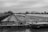 A black and white image featuring a large river barge laden with gravel or sand, positioned on a calm waterway. The barge is flanked by a metal siding on one side and trees on the other. The overcast sky adds a moody atmosphere to the industrial scene. In the background, there are bridges and a few buildings visible across the landscape.