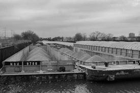 A black and white image featuring a large river barge laden with gravel or sand, positioned on a calm waterway. The barge is flanked by a metal siding on one side and trees on the other. The overcast sky adds a moody atmosphere to the industrial scene. In the background, there are bridges and a few buildings visible across the landscape.