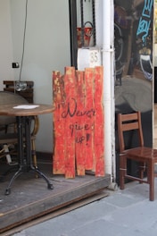 A wooden sign painted in red with the motivational message 'Never give up!' leans against the wall outside a café. The setting includes a wooden table and chair on a raised platform. Part of a black reflective wall and a door with the number 35 are visible.