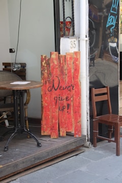 A wooden sign painted in red with the motivational message 'Never give up!' leans against the wall outside a café. The setting includes a wooden table and chair on a raised platform. Part of a black reflective wall and a door with the number 35 are visible.
