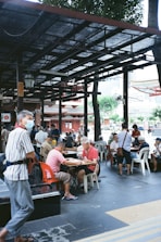 People are gathered outdoors under a sheltered area with open sides, sitting on plastic chairs and playing board games at several tables. The environment seems relaxed, with trees and a building visible in the background. A few individuals are standing or walking nearby, and most appear to be older adults.