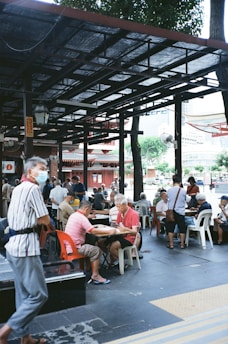 People are gathered outdoors under a sheltered area with open sides, sitting on plastic chairs and playing board games at several tables. The environment seems relaxed, with trees and a building visible in the background. A few individuals are standing or walking nearby, and most appear to be older adults.