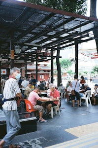 People are gathered outdoors under a sheltered area with open sides, sitting on plastic chairs and playing board games at several tables. The environment seems relaxed, with trees and a building visible in the background. A few individuals are standing or walking nearby, and most appear to be older adults.