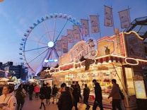 A bustling fairground scene with a large ferris wheel illuminated in blue and pink lights. In the foreground, there is a vibrant market stall with bright signage offering various sweets and treats. Many people are walking around, some holding food and drinks. Numerous flags are displayed above the stall, adding to the lively atmosphere.
