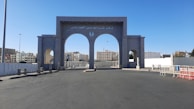 An open plaza features an impressive, large, gray archway with three arched openings. The central arch is larger and contains Arabic writing at the top. Behind the archway, modern buildings are visible under a clear blue sky. Barricades and streetlights are also present, contributing to the urban environment.