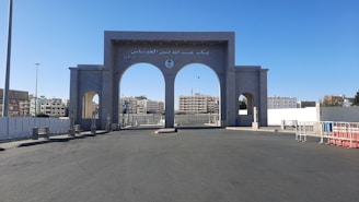 An open plaza features an impressive, large, gray archway with three arched openings. The central arch is larger and contains Arabic writing at the top. Behind the archway, modern buildings are visible under a clear blue sky. Barricades and streetlights are also present, contributing to the urban environment.
