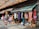 A vibrant marketplace stall is filled with colorful hanging garments, bags, and various items for sale. Mannequins display clothing on the sidewalk, and sunlight creates patterned shadows through a lattice above.