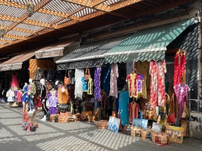 A vibrant marketplace stall is filled with colorful hanging garments, bags, and various items for sale. Mannequins display clothing on the sidewalk, and sunlight creates patterned shadows through a lattice above.