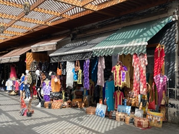 A vibrant marketplace stall is filled with colorful hanging garments, bags, and various items for sale. Mannequins display clothing on the sidewalk, and sunlight creates patterned shadows through a lattice above.