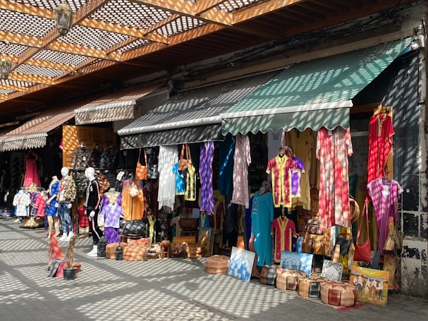 A vibrant marketplace stall is filled with colorful hanging garments, bags, and various items for sale. Mannequins display clothing on the sidewalk, and sunlight creates patterned shadows through a lattice above.