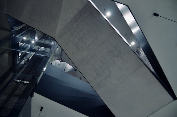A modern architectural interior featuring geometric shapes and concrete surfaces. The space includes large reflective glass panels and sharp angles with diffused lighting creating a stark, minimalist aesthetic.