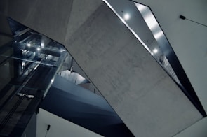A modern architectural interior featuring geometric shapes and concrete surfaces. The space includes large reflective glass panels and sharp angles with diffused lighting creating a stark, minimalist aesthetic.
