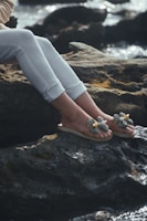 A person's legs and feet wearing decorative sandals are resting on rocky terrain by the water's edge. The person is wearing white pants, and the focus is on the detailed design of the sandals, which have floral decorations.